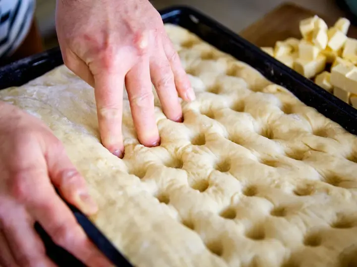Hände drücken Vertiefungen in den Teig für ein Butterkuchen Rezept. Würfel aus Butter liegen bereit.