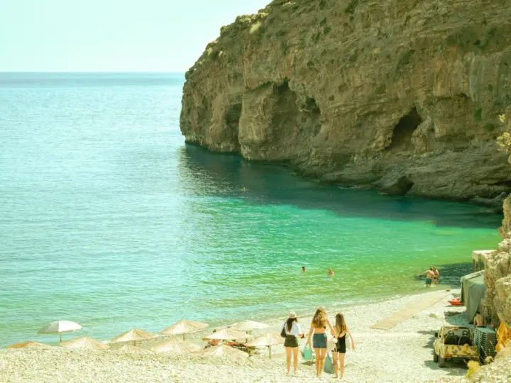 Trzy dziewczyny idące po kamienistej plaży w kierunku turkusowego morza przy skalistym klifie.