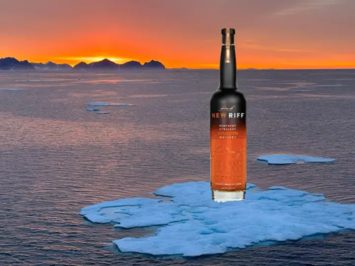 Eine Flasche New Riff Bourbon Whiskey steht auf einer Eisscholle im Meer. Der Sonnenuntergang taucht die Berge am Horizont in warmes Licht. Ein Moment der Ruhe für den besten Whisky der Welt.