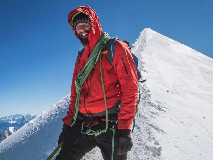 Uśmiechnięty alpinista na szczycie Mont Blanc, w czerwonej kurtce i z liną, gotowy na dalszą wspinaczkę.
