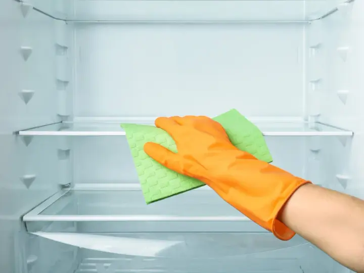 Main d'une personne portant un gant orange, essuyant une étagère de réfrigérateur vide avec une éponge verte. Le nettoyage du frigo est terminé.