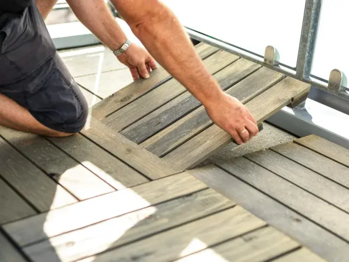 Un homme installe des dalles de terrasse en bois sur son balcon. Une belle idée sol balcon pour un espace extérieur accueillant.
