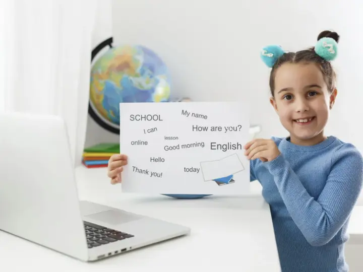 Uśmiechnięta 6latka z tabliczką "English" i zwrotami do nauki. Obok laptop i globus.