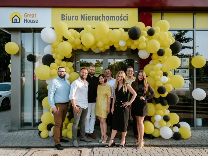 Grupa ludzi pozuje przed biurem nieruchomości ozdobionym żółtymi balonami.