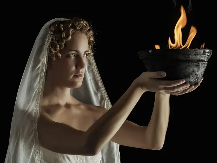 A woman with curly hair and a veil, embodying Hestia goddess, holds a bowl with a sacred flame.