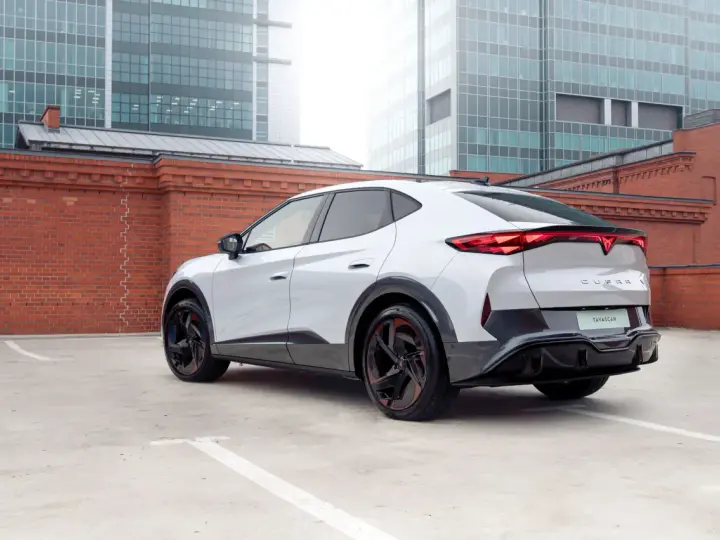 Biały elektryczny SUV Cupra Tavascan z czerwonymi akcentami na felgach, zaparkowany na tle ceglanego muru i nowoczesnych budynków.