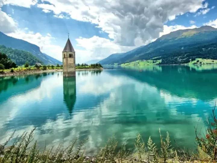 Il lago del campanile, con la sua torre sommersa, riflette montagne verdi e un cielo nuvoloso.