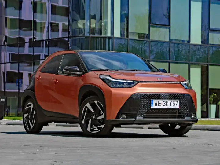 Pomarańczowy Toyota Aygo X Hybrid z czarnymi detalami stoi przed nowoczesnym budynkiem.