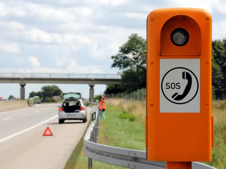 Ihr Fahrzeug ist auf der Autobahn liegen geblieben. Eine Frau in Warnweste steht neben dem offenen Kofferraum, ein Warndreieck liegt auf der Fahrbahn. Eine Notrufsäule steht im Vordergrund.