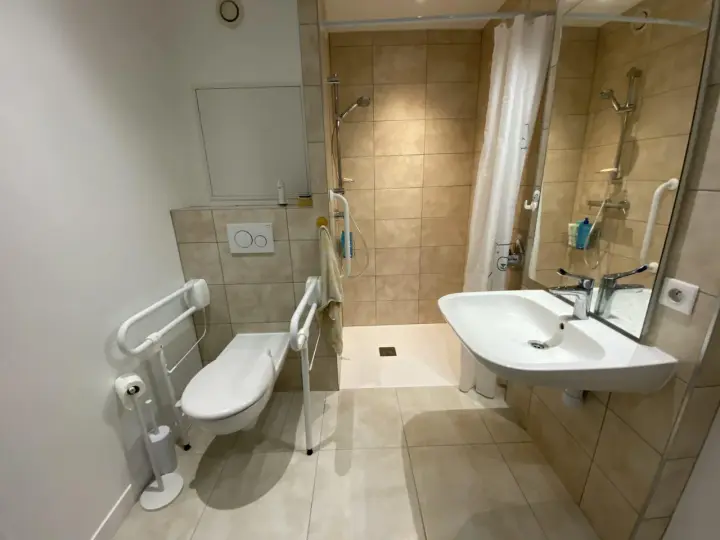 Salle de bain pmr avec douche à l'italienne, WC avec barres d'appui et lavabo. Confort et sécurité pour tous.