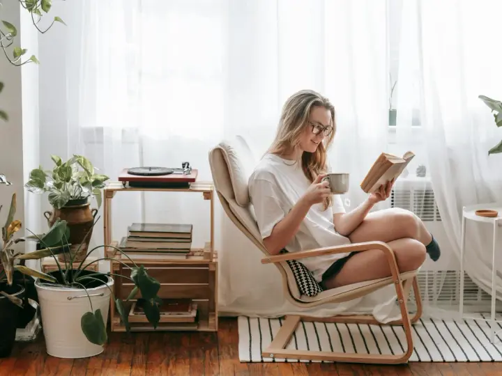Mujer con gafas disfruta de un libro y una taza en su rincón de lectura, rodeada de plantas y luz natural.