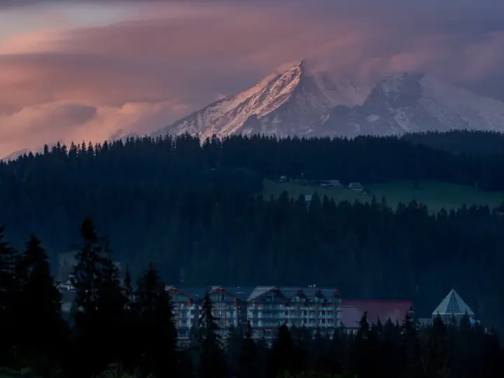 Górski krajobraz o zachodzie słońca z ośnieżonymi szczytami, lasem i budynkami hotelowymi.