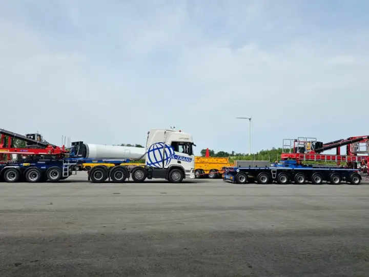 Ciężarówka z naczepą tandem przewozi element turbiny wiatrowej. Obok stoi inna naczepa z dźwigiem.