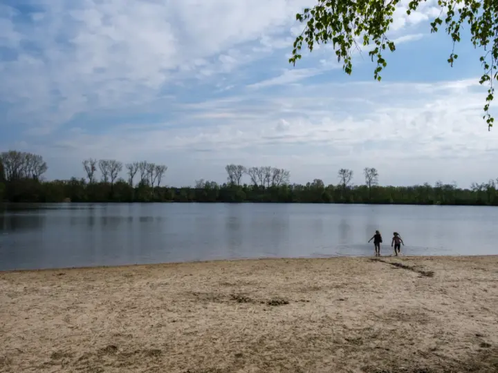 Zwei Kinder planschen im Wasser am Sandstrand des Dieksees in Lingen. Bäume säumen das Ufer unter einem bewölkten Himmel.