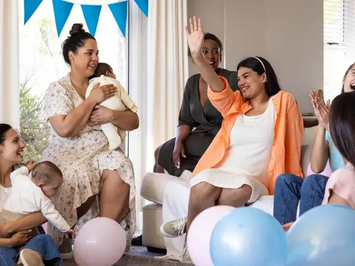 Glada kvinnor firar en babyshower med ballonger och bebisar. En kvinna vinkar glatt.