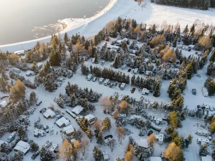 Wintercamping im Erzgebirge: Wohnmobile und Wohnwagen stehen im Schnee auf einem Campingplatz am See, umgeben von verschneiten Bäumen.