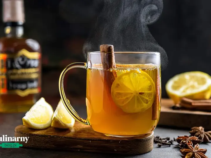 Gorąca herbata z whisky, cytryną i cynamonem, parująca w szklance. Obok leżą cytryny i przyprawy.