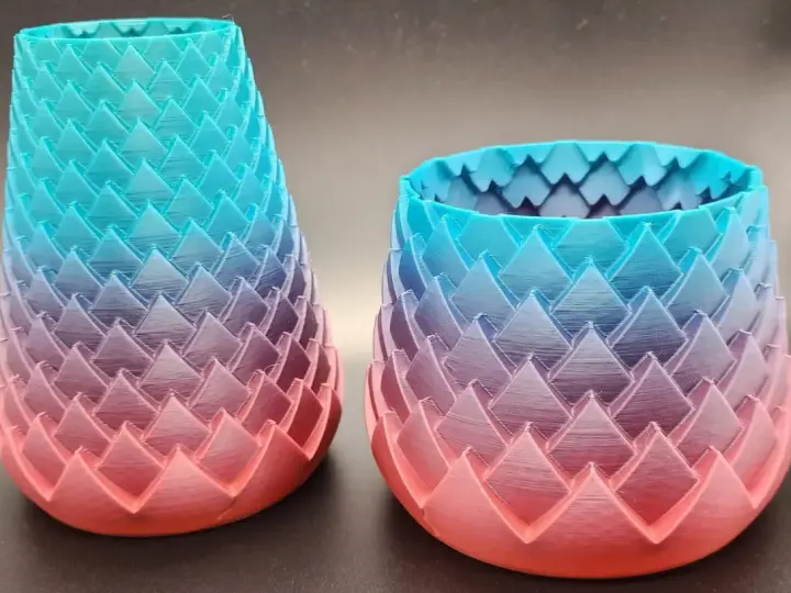 Two dragon-scale patterned vases, created with color 3d printing, showcase a gradient from coral pink to teal blue.