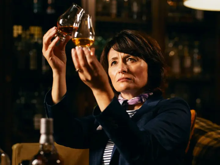 Rachel Barrie betrachtet konzentriert zwei Gläser Whisky, während sie in einer dunklen Bar sitzt.