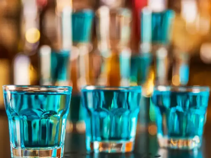 Trzy szklanki z niebieskim płynem, gotowe do wypicia jako kamikaze shot. W tle bar z butelkami.