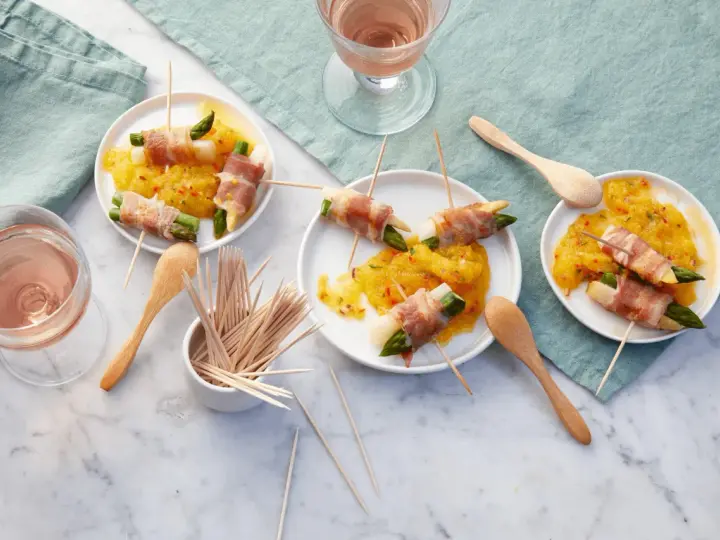 Leckere Spargel im Speckmantel auf gelbem Chutney, serviert auf kleinen Tellern mit Holzspießen und Roséwein.
