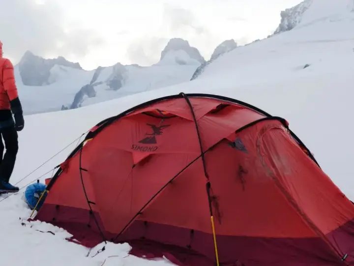Alpinista w czerwonej kurtce stoi obok namiotu na tle ośnieżonych szczytów. W oddali widać groźne góry, gdzie mogą wystąpić lawiny w Alpach.