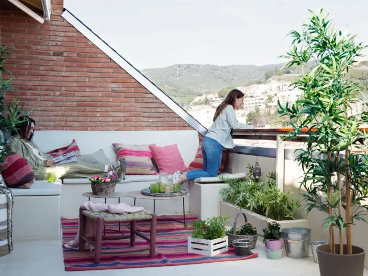 Dos mujeres disfrutan de una terraza con arboles bonitos, cojines coloridos y vistas a la montaña.
