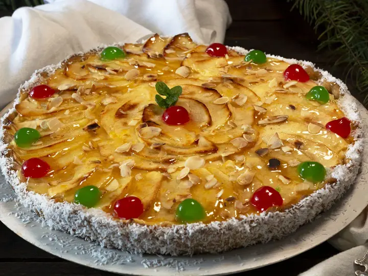 Deliciosa tarta de manzana cubierta de almendras laminadas y cerezas rojas y verdes. El borde está cubierto de coco rallado.