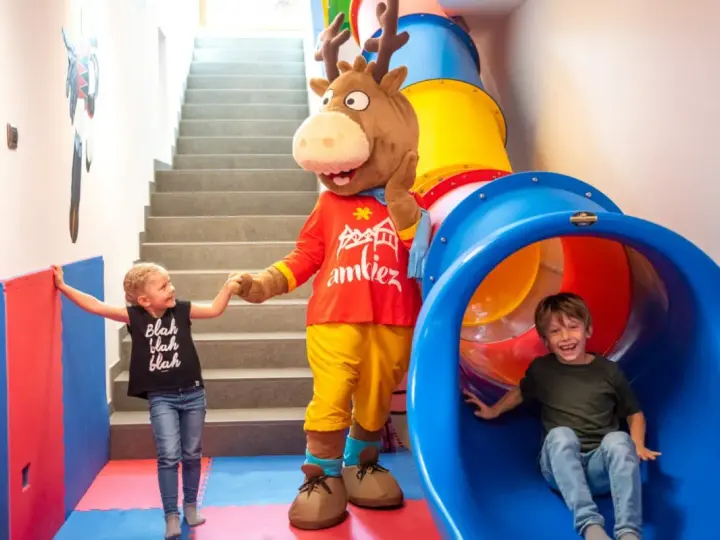 Mascot and two children in a colorful indoor playground with a slide and stairs.