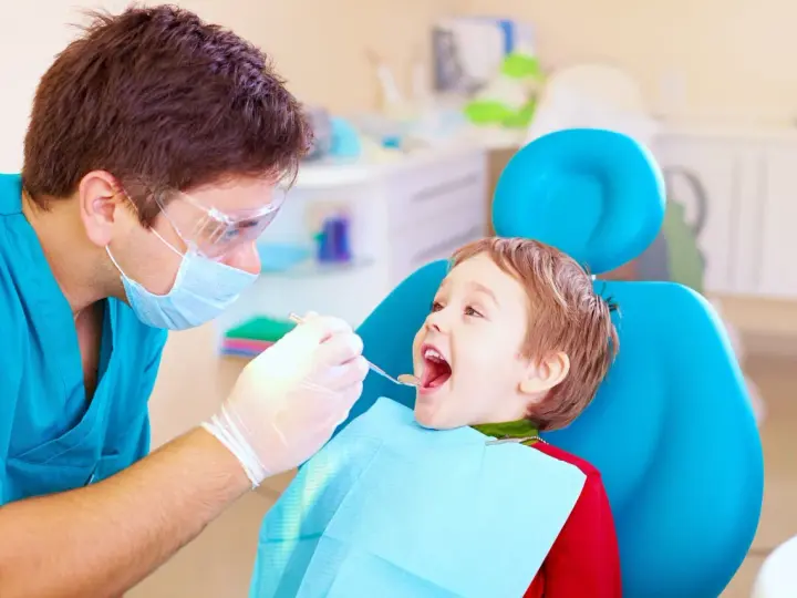 Dentysta bada uśmiechniętego chłopca, sprawdzając, kiedy najpóźniej pierwszy ząbek pojawi się u malucha.