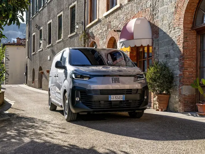 En grå Fiat Doblo står parkerad på en smal gata i Italien. En vacker byggnad med tegel och sten syns i bakgrunden.