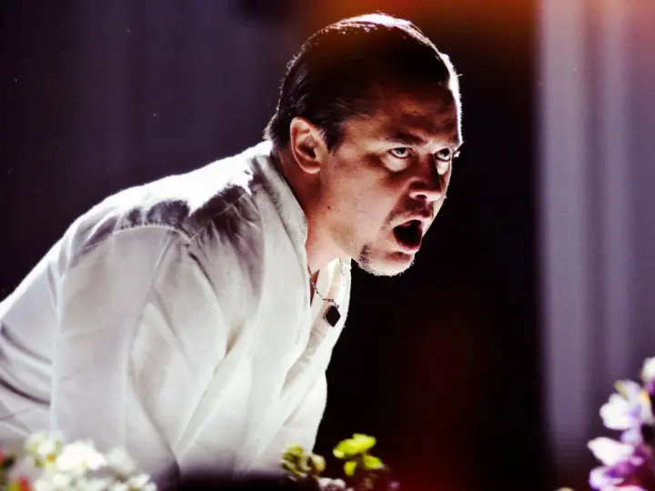 Mike Patton heute, mit weit aufgerissenem Mund und intensivem Blick, trägt ein weißes Hemd. Blumen im Vordergrund.