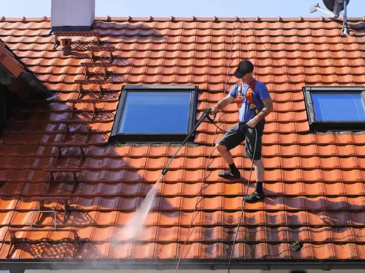 Un homme effectue le nettoyage toiture avec un nettoyeur haute pression sur un toit en tuiles rouges, sous un ciel bleu.