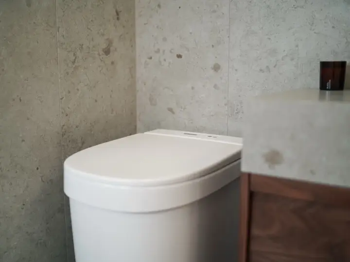 Modernt badrum med en vit toalett utan avlopp, grå kaklade väggar och ett handfat i betong.