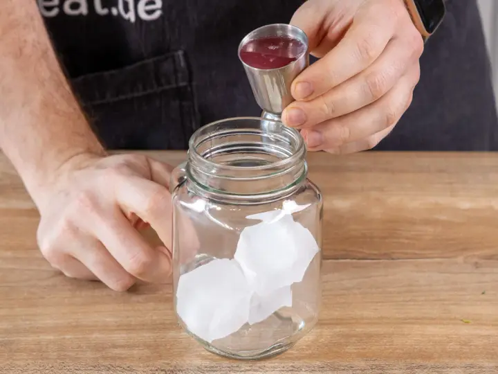 Eiswürfel in einem Glas, dazu wird rote Flüssigkeit aus einem Messbecher gegossen. So einfach lässt sich Rhabarberschorle selber machen!