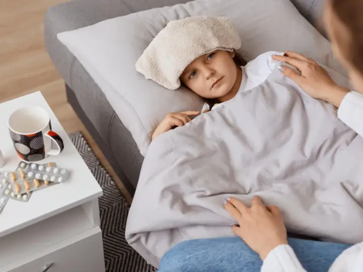 Chora dziewczynka z okładem na głowie leży pod kocem. Obok leki i kubek. Mama czuwa, zapewniając opiekę na dziecko.
