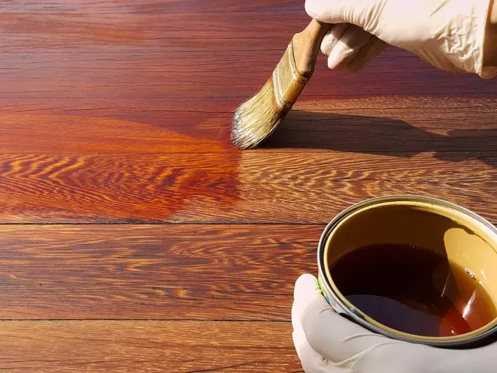 Mano guantata applica una vernice scura su assi di legno, mostrando come scurire il legno per un aspetto più profondo e ricco.