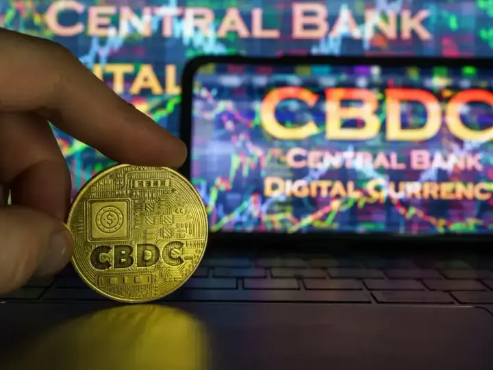 Złoty żeton z napisem CBDC w dłoni, na tle ekranu z wykresem i napisem "CENTRAL BANK DIGITAL CURRENCY". Potencjalne zagrożenia CBDC.