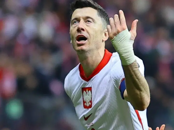 Robert Lewandowski w koszulce reprezentacji Polski, z bandażem na ręce, wznosi gest ku górze. Fani zastanawiają się, o której mecz piłki nożnej.