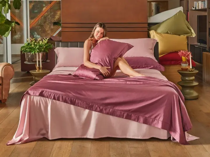 Una donna sorride su un letto con lenzuola rosa. Per mantenerle fresche, si lavano a 40 gradi.