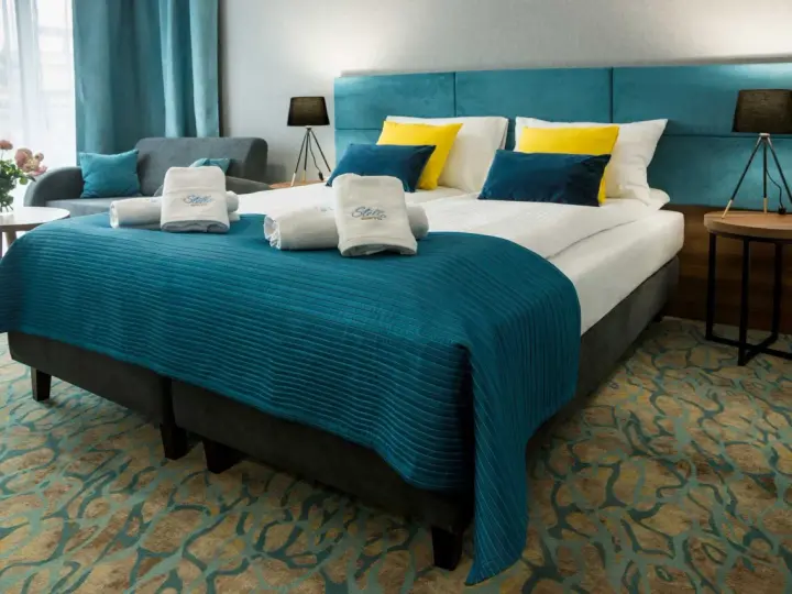 Przytulny pokój hotelowy z dużym łóżkiem, ozdobionym turkusową narzutą i żółto-niebieskimi poduszkami. Ręczniki z logo Stella Resort & Spa zachęcają do relaksu.