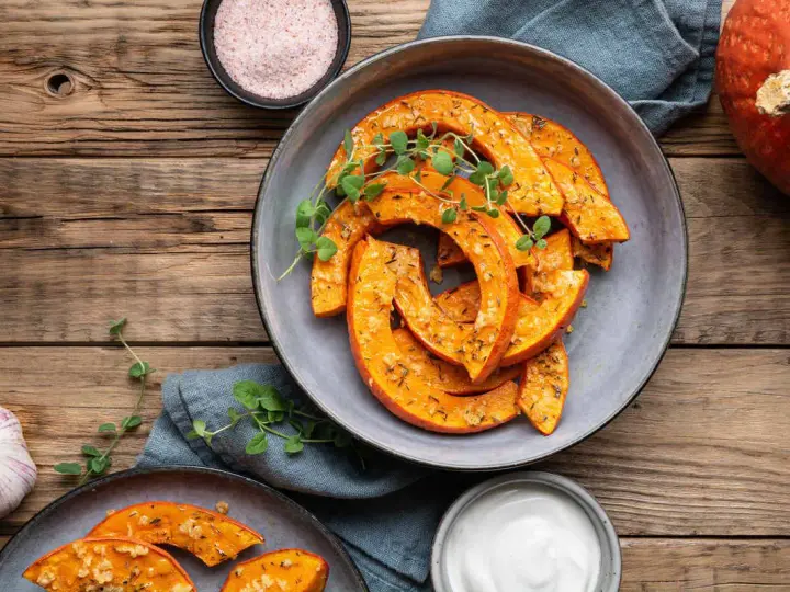 Fette di zucca in agrodolce in padella, condite con erbe aromatiche e servite su un piatto grigio, con contorno di aglio e sale rosa.
