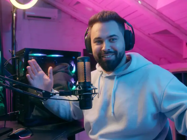 Uśmiechnięty streamer w słuchawkach, przy mikrofonie, tłumaczy, streaming co to jest. W tle komputer i różowe oświetlenie.