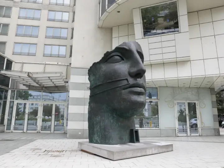 Okaleczona, ciemna rzeźba głowy z opaską na ustach, na tle nowoczesnego budynku.