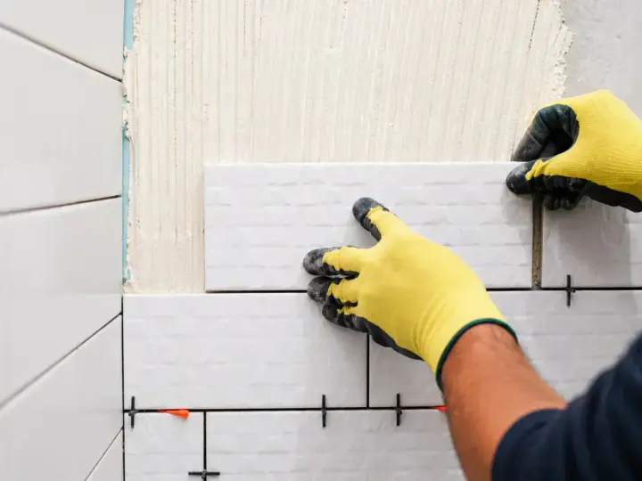 Mains gantées de jaune en train de coller du carrelage sur du carrelage, avec des croisillons pour un espacement parfait.