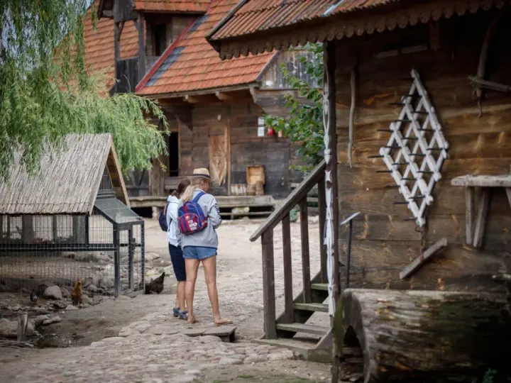 Dwie dziewczyny zwiedzają skansen, który może być częścią oferty hotelu bon turystyczny. W tle drewniane chaty i kurnik.
