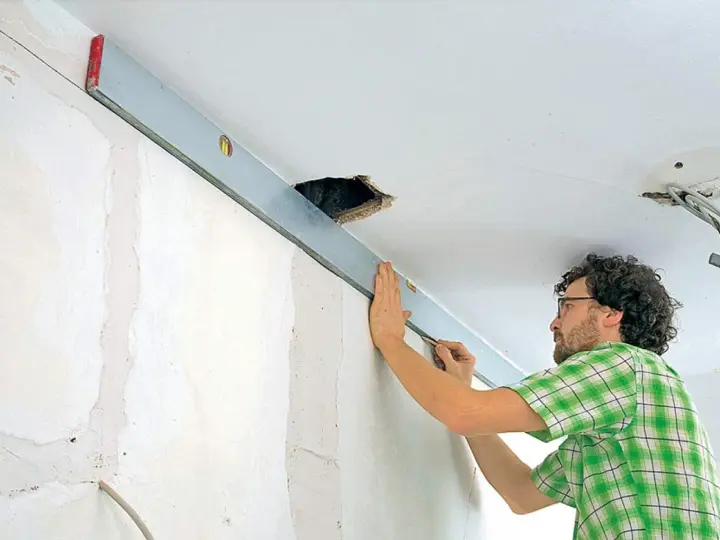 Mann richtet eine Wasserwaage an der Decke aus, um die Wand mit Rigips zu verkleiden. Kabel hängen aus einer Deckenöffnung.