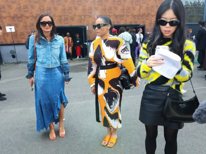 Tres mujeres posan con atuendos de moda. La del centro lleva un vestido estampado y sandalias naranjas, un ejemplo de que son mules.