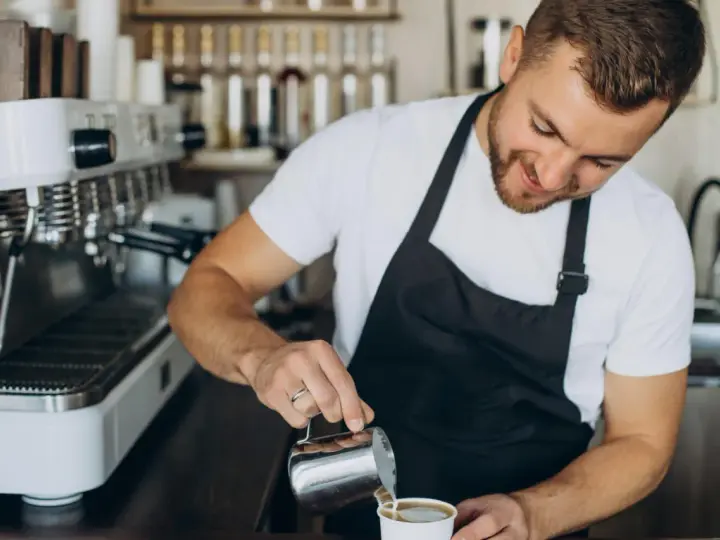 Uśmiechnięty barista w czarnym fartuchu nalewa mleko do kawy. Obok stoi profesjonalna ekspres do kawy.