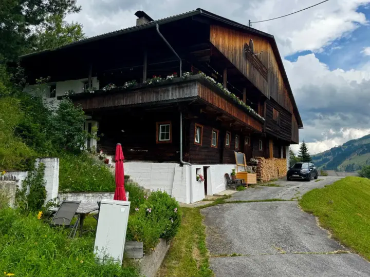 Drewniana austeria w górach, z balkonami ozdobionymi kwiatami. Obok stoi czarny samochód, a w oddali widać zalesione wzgórza.